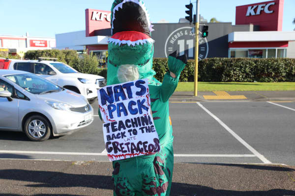 A teacher dressed in a dinosaur costume holding a sign during a protest in West Auckland