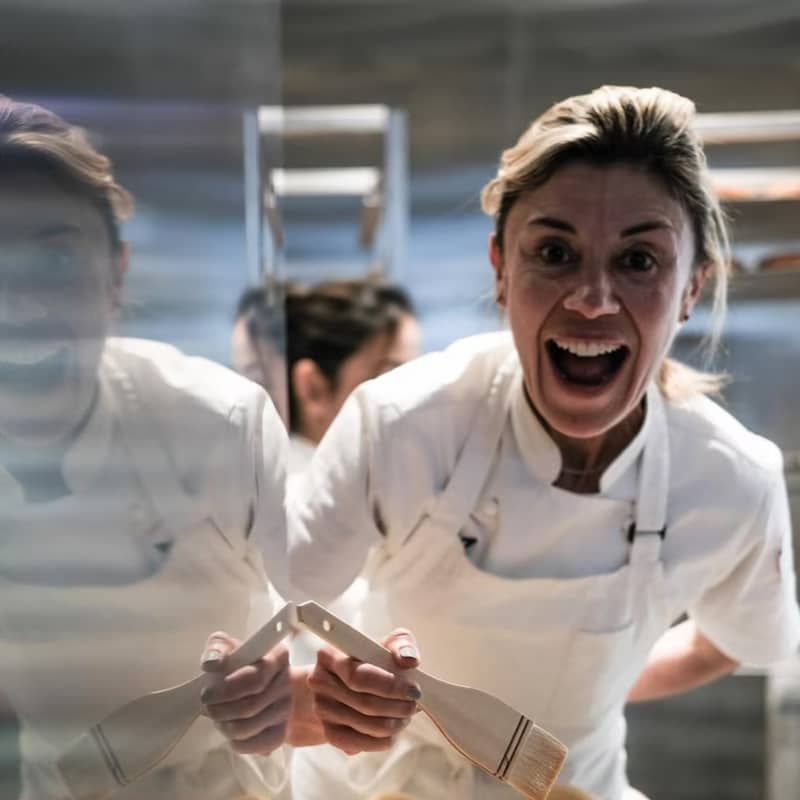 A smiling woman in a white chef's outfit paints uncooked croissants with a brush.