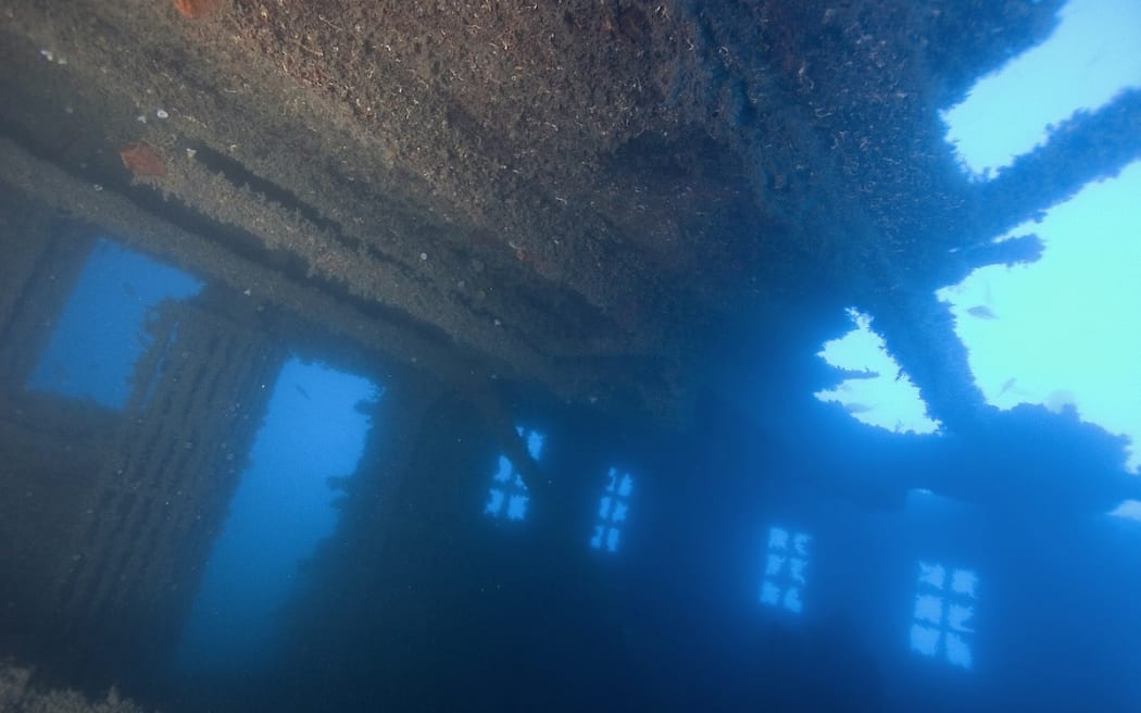 The inside of a once-luxurious library of the RMS Niagara. The roof has gaping holes and the structure is covered in rust and growths. Fish swim in and around the room.