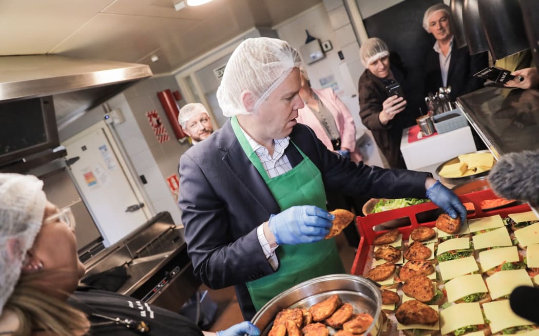 Chris Hipkins is visiting the Post Office Hotel in Pahiatua. They make free school lunches for approx 900 students in the area. Hipkins is helping make burgers which will be delivered to schools later.