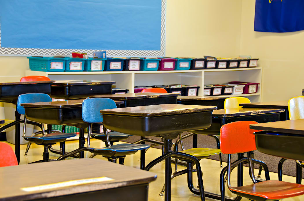 Students away from a classroom, which is filled with empty school desks.