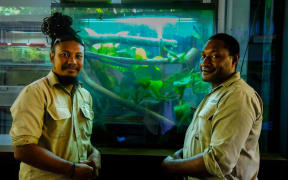 Ryan and his colleague, from Papua New Guinea, stand in front of a tank filled with greenery. They are both wearing khaki shirts.