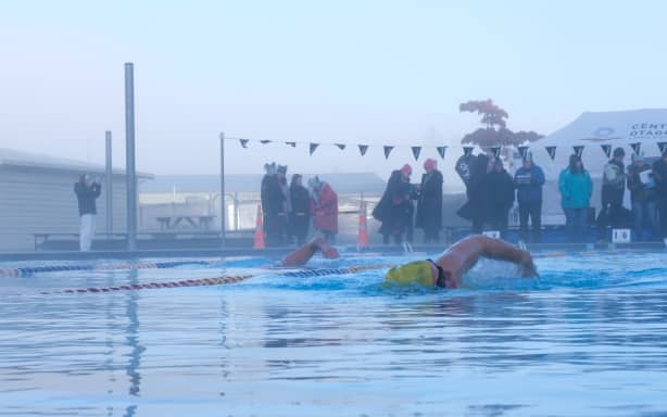 A swimmer in a yellow cap and black goggles in the centre of the picture swims ahead of another swimmer to her left. It's an outdoor pool with low morning light and morning mist. Some supporters in jackets and hats can be seen at the end of the pool.