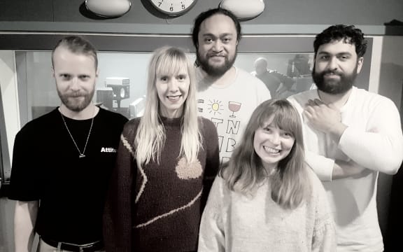 Five band members standing in the RNZ recording studio against the control room window