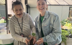 Shirleen and Nic smile broadly. Shirleen is holding a trowel and seedling.