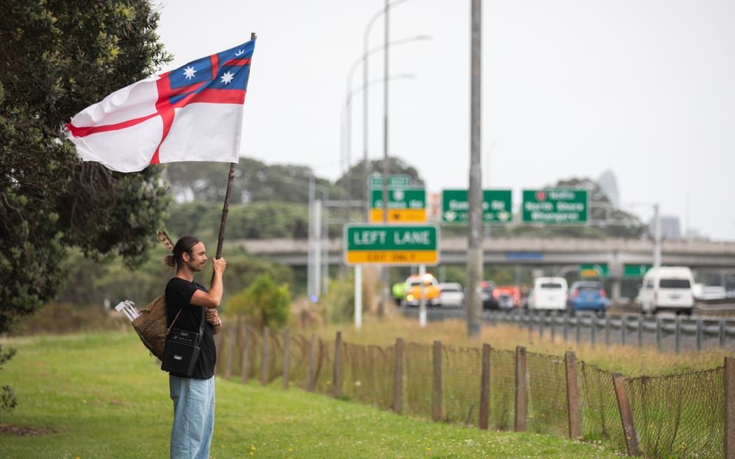 A participant in Hikoi mō Te Tiriti stands near State Highway One before the group crosses the Habour Bridge holding the United Tribes of New Zeland flag.