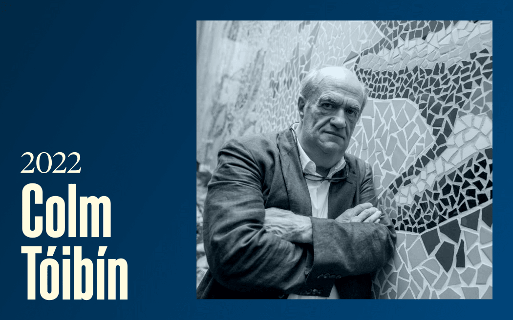 A slightly frowning man stands with his arms crossed in front of a mosaic wall, text reads "2022, Colm Tóibín"