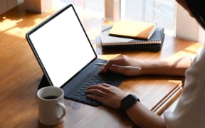 Cropped image of young beautiful woman working as writer typing on computer tablet with white blank screen while sitting at the wooden working table with sunlight through windows as background.