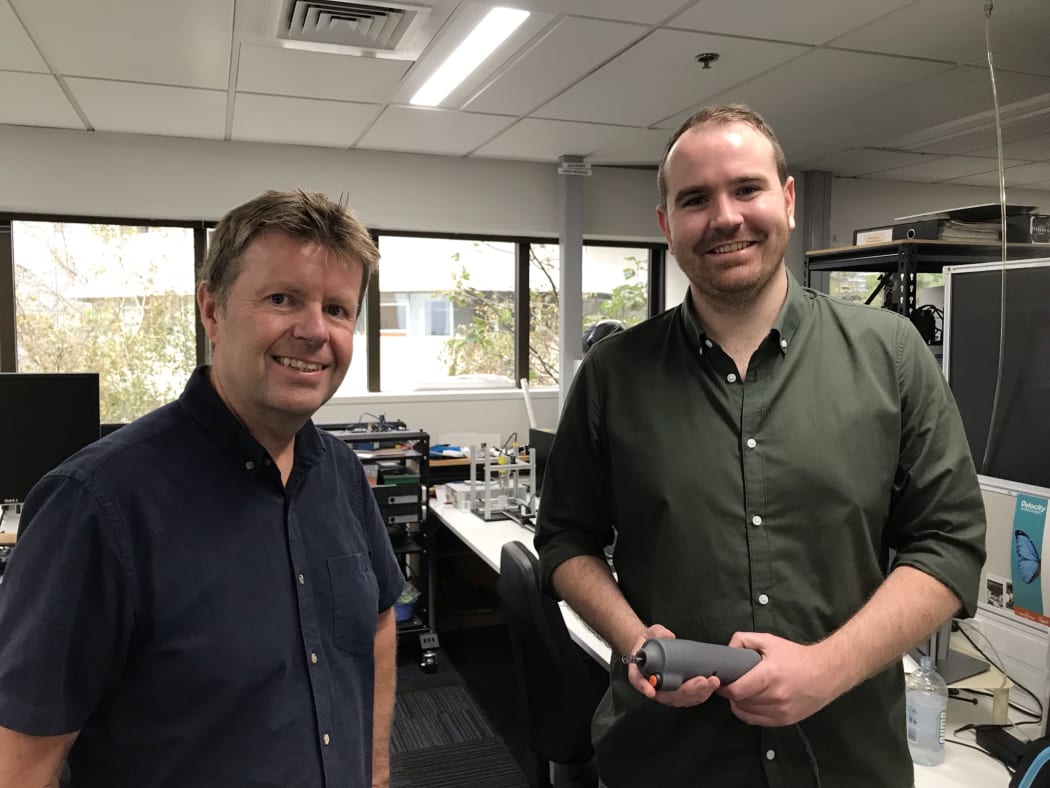 Andrew and James stand next to each other, James has the research version of the electric motor jet-injector in his hand - its a cylindrical grey shape.