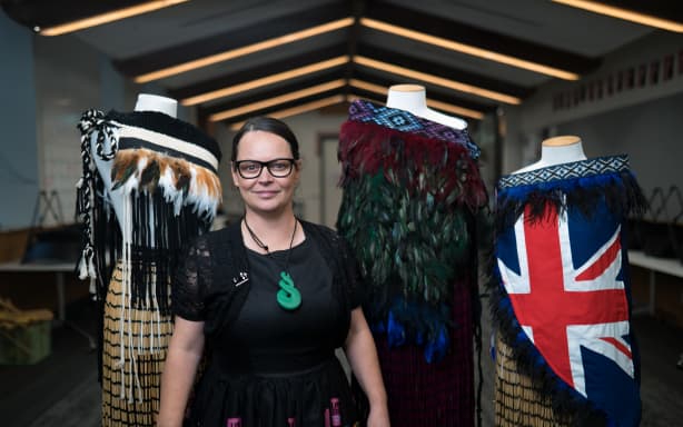 A portrait of Nan in a large hall space in front of three headless mannequins with korowai on them. She is wearing a 3D printed green Pikorua.