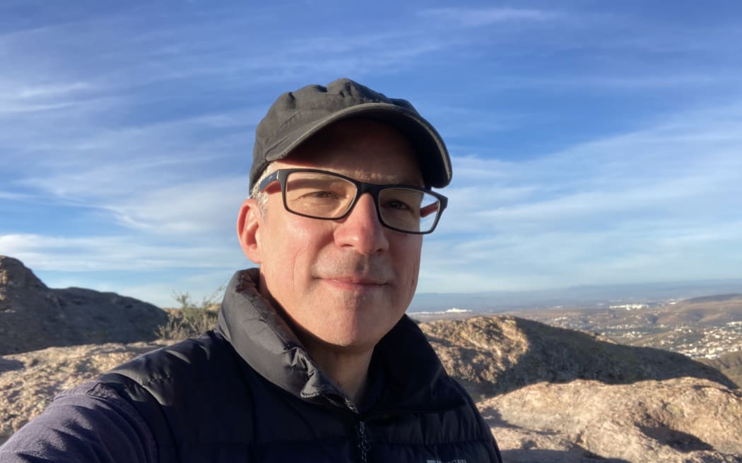 Étienne smiles at the camera. He is wearing a puffer jacket and stands atop a mountain under a crisp blue sky.