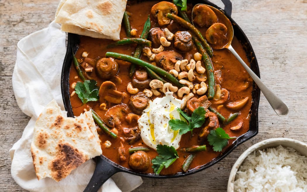 A cast iron frying pan filled with a rich red-brown curry of mushrooms, cashews and green beans, garnished with flatbreads, coriander and yoghurt.