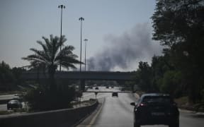 Motorists drive along a street as smoke rises from a reported Iranian strike in the area where the US Embassy is located in Kuwait City on March 2, 2026. Black smoke was seen rising from the US embassy in Kuwait City on March 2 after the latest volley of Iranian strikes, an AFP correspondent saw. (Photo by AFP)