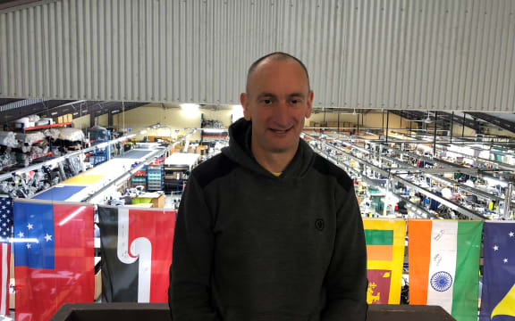 Ben Kepes wears a black hoodie and stands on an elevated platform. Behind him is the factory floor of Albion Clothing, with rows of workbenches and tools. There is a string of flags hanging over the factory floor.