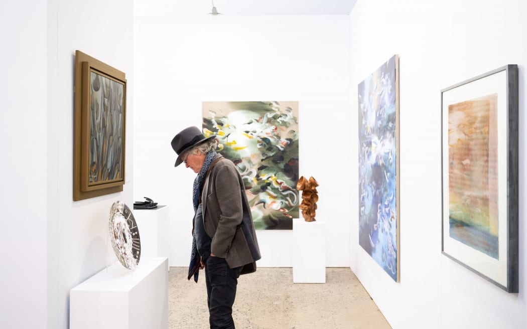 A man in a tweed jacket and hat looks at objects in a well-lit gallery space.