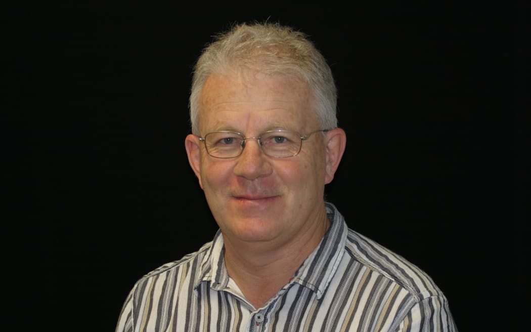 Rod Thomas smiles at the camera. He is wearing a striped, collared shirt and glasses. He is set against a black backdrop.