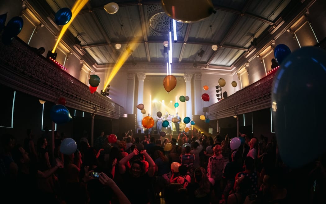 A dance hall lit up with balloons and strobe lights. People dance while a DJ plays on stage.
