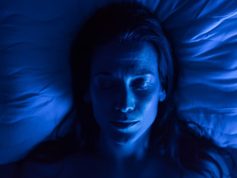 A woman lies with closed eyes on a pillow in blue light.