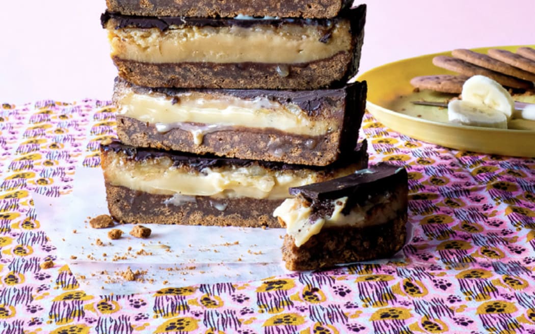 A vertical stack of five pieces of banana caramel slice, showing a cross-section of biscuit base, gooey caramel and chocolate topping, on a pink patterned cloth.