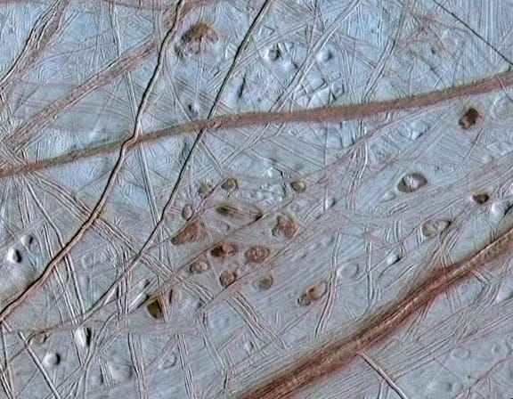 Reddish spots and shallow pits peppering the enigmatic ridged surface of Jupiter's moon Europa.