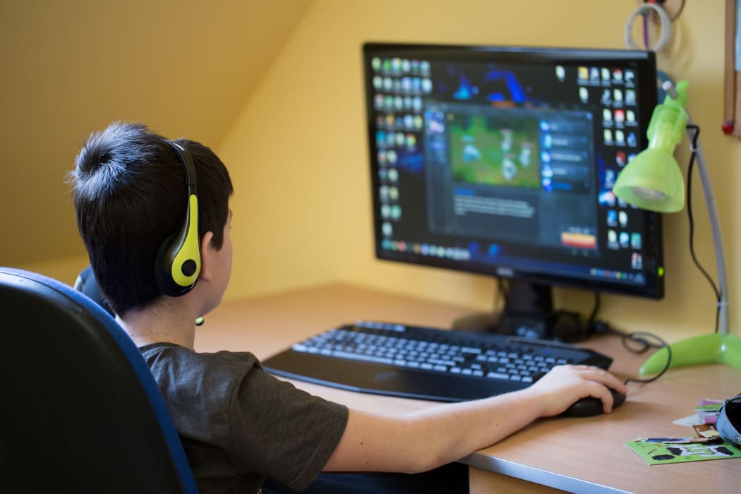 A photo of a teenager boy taken from behind. He is using computer at home with headphones to play a game in his room.