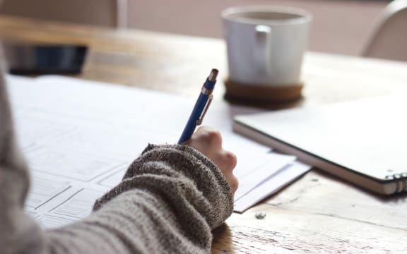 A hand holding a pen hovers over paper with a mug in the background