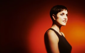 Princess Chelsea wears a black dress and stands in front of an redish orange backdrop. She is lit with a spotlight and had a 1920s sort of aesthetic.