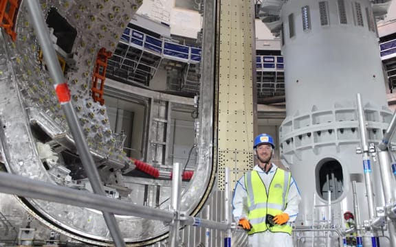 Dr Tom Wauters, smiling and wearing a hard hat and high vis, stands in front of a large D-shaped module.
