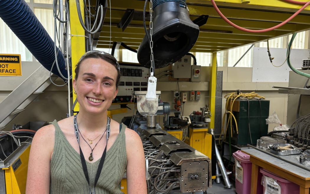 Louise stands in front of a large yellow metal frame under which is a big black extract hood and a long metal rectangle with lots of metal wires on the left hand side. She is smiling, with her hair tied back, wearing a black lanyard a green sleeveless top and a pounamu necklace.