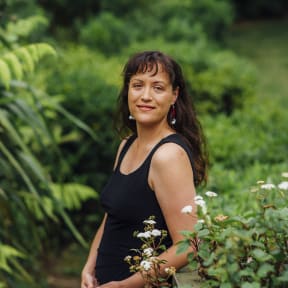 Māori architect Jade Kake wears a black tank top and pants and stands in front of greenery outside.