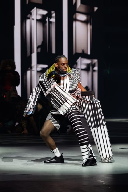 A black man crouches in a striped black and white garment which resembles cloth armour.