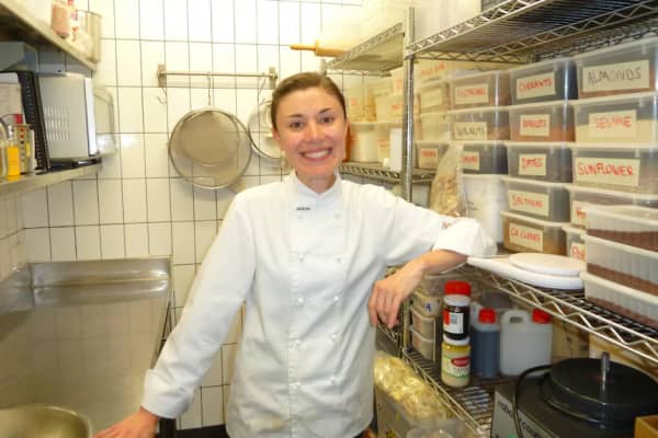 A smiling woman in a white jacket stands in a pantry.