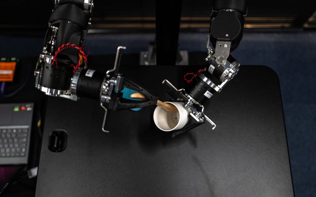 A pair of swivelling, human-like robotic arms, built for physical artificial intelligence research, mirror the motions of an operator in a VR headset twirling his hands like a magician.