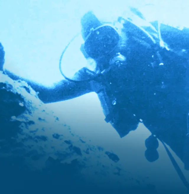 Blue-tinted underwater photo showing a scuba diver in full gear holding onto a large rock or underwater structure. The diver appears to be inspecting or working on something submerged. The lighting gives the image a dramatic, archival feel.