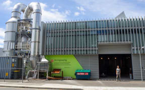 A woman stands in the open garage door of a large warehouse-style building, with big steel pipes extending from its roof. The building is labelled 'Fire engineering'.