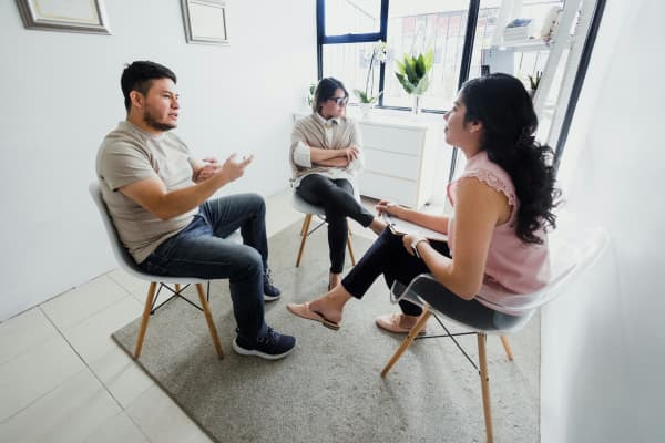 Couple sitting in separate chairs speaking with a woman holding a clipboard.