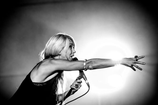 In a monochrome photograph, a woman with long blonde hair sings into a microphone with her other arm outstretched.