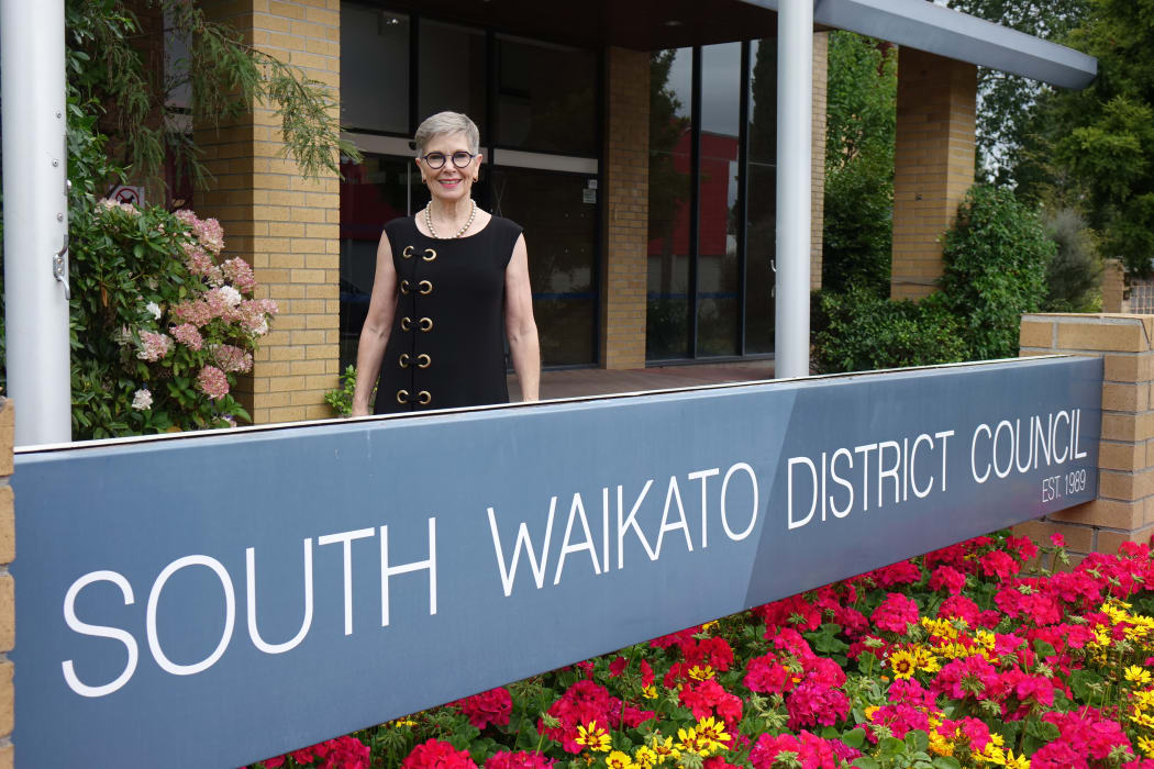 Female mayor stands behind a council sign with bright municpal flowers planted in front