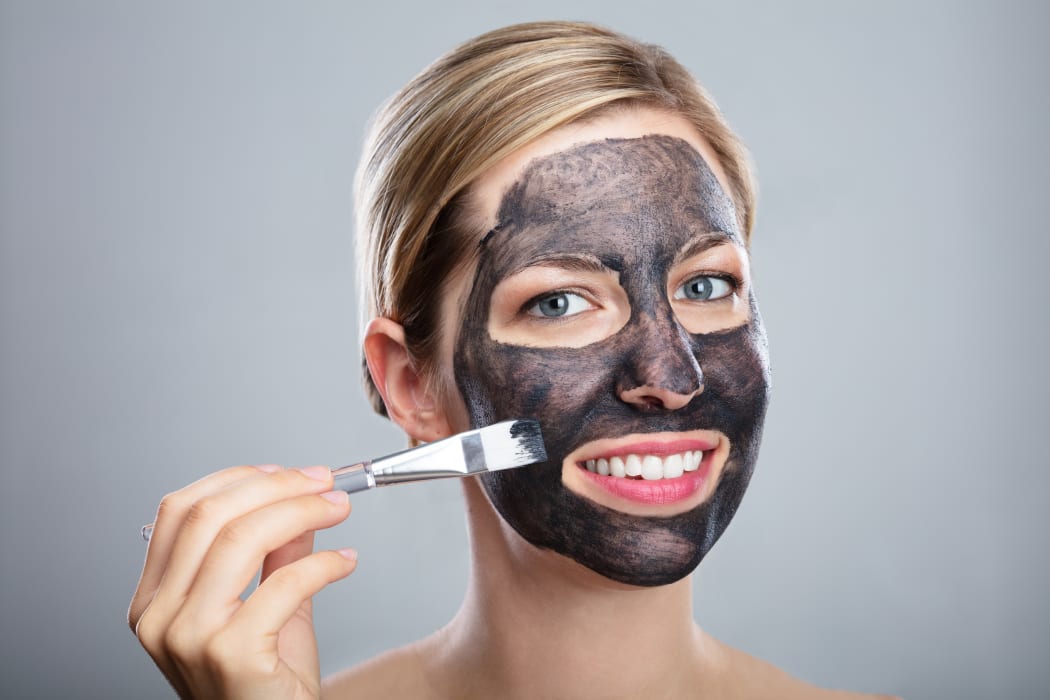 Young Woman Applying Activated Charcoal Face Mask With Brush On Grey Background