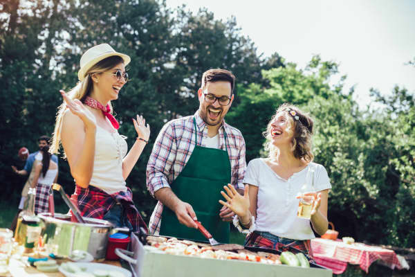 Group of people standing around grill, chatting, drinking and eating.