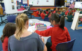 A view from behind of teacher aide and two little girls while rest of class taught  further into classroom.