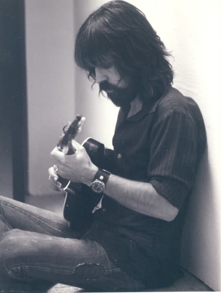 Clarence White plays an accoustic guitar while wearing a dark shirt and standing against a white wall.