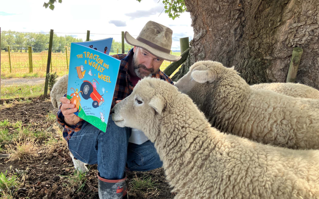 Tim is holding a copy of his children's book 'The tractor Has A Wobbly Wheel', and reading it to a couple of sheep.
