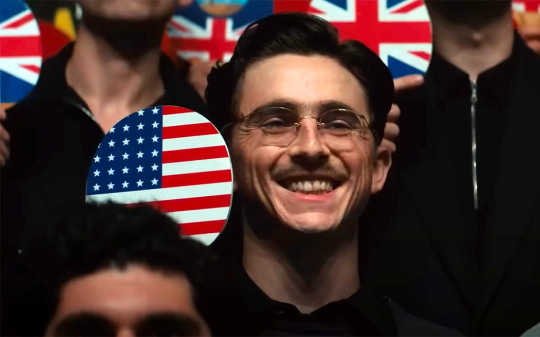 As Marty Supreme, Timothée Chalamet is a beaming young man holding up a ping-pong racquet with the American flag.