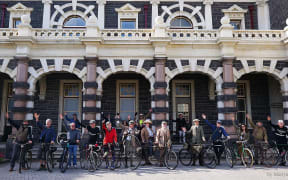 A group of people pose on their bikes in front of the Dunedin Railway Station. Many are dressed in tweed.