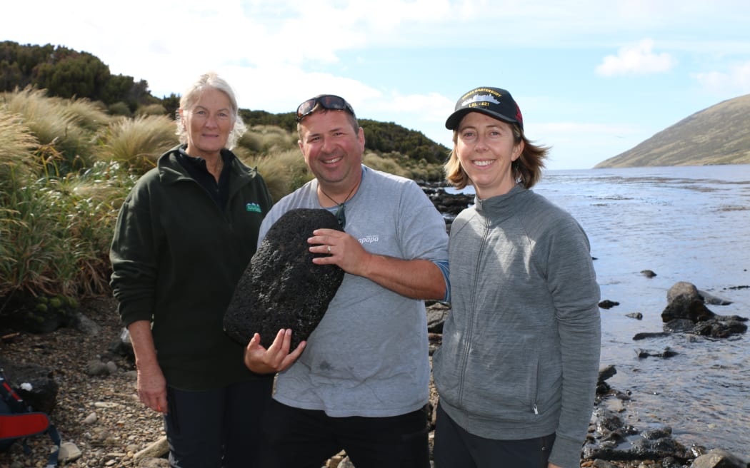 The three people are smiling in the sunshine, they are on a stony beach beside the water. Bob, in the middle holds a large rock to his chest.