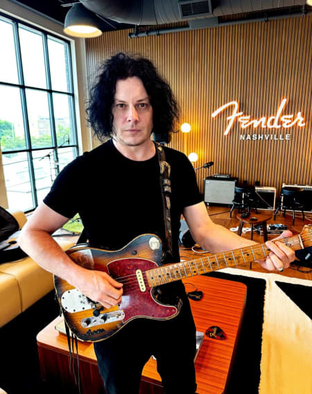 Jack White - wearing a black t-shirt - holds a guitar in front of a neon sign that says the word 'Fender'.
