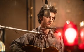 Film still of actor Timothée Chalamet playing Dylan, performing on guitar and harmonica