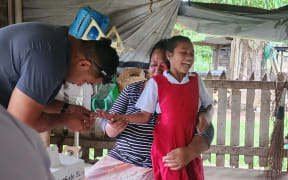 Young girl in red school pinafore gets tested for Hepatitis  in outdoor room sheltered by tarpaulins