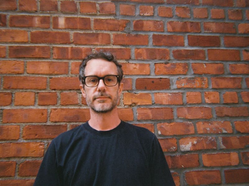 Tony Stamp is a slightly smiling man with glasses and a beard, wearing a black sweater and standing against a brown brick wall.